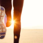 La imagen muestra un primer plano de una persona corriendo sobre una superficie arenosa, probablemente una playa, con el sol brillando intensamente en el fondo.