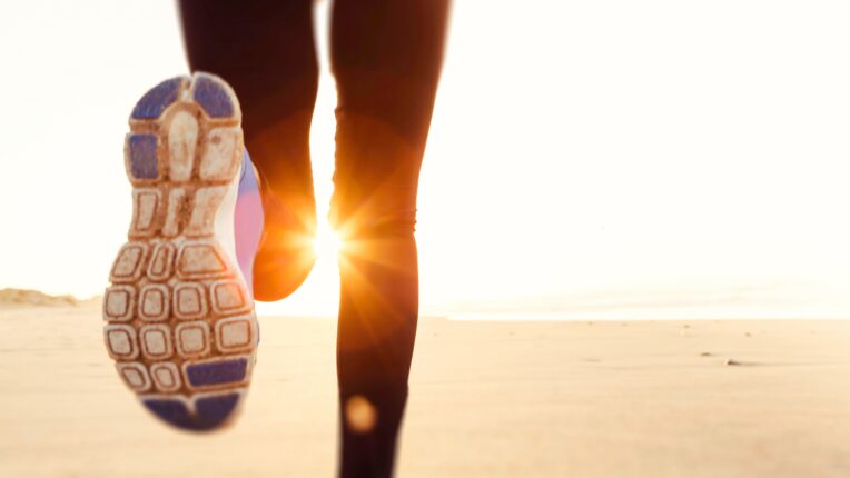 La imagen muestra un primer plano de una persona corriendo sobre una superficie arenosa, probablemente una playa, con el sol brillando intensamente en el fondo.