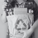 La imagen muestra a una persona sosteniendo una bolsa de papel con el símbolo de reciclaje impreso en ella. Dentro de la bolsa hay varios artículos reciclables, incluyendo una botella de plástico y una caja de cartón. La persona lleva una camisa de cuadros y tiene las uñas pintadas de un color oscuro