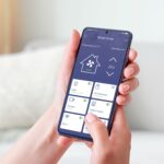 Mano sosteniendo un teléfono inteligente que tiene abierta una aplicación de "Smart Home". La pantalla de la aplicación exhibe varios controles y lecturas relacionados con la automatización del hogar.